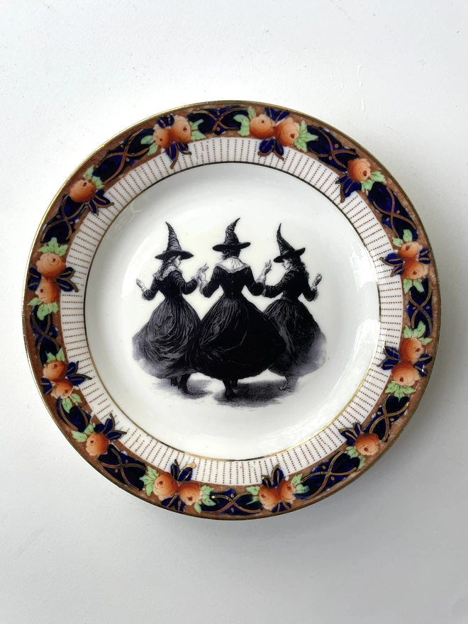 a coven of 3 witches dance hand in hand, the image is victorian in black and white and is printed directly onto an ornate vintage plate
