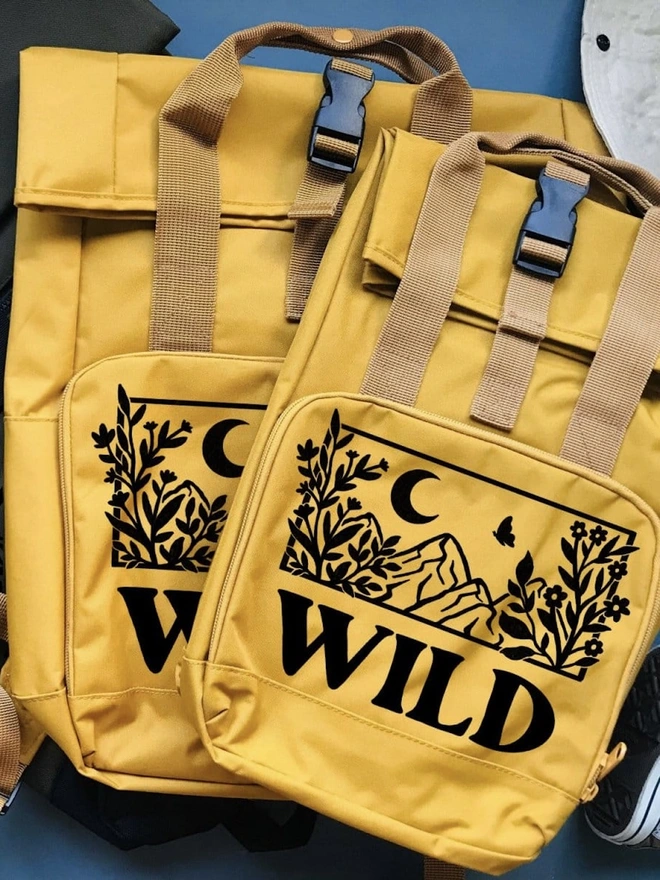 'Wildings' Children’s Recycled Roll Top Back Pack, a pair of colourful backpacks sitting on a blue surface. 