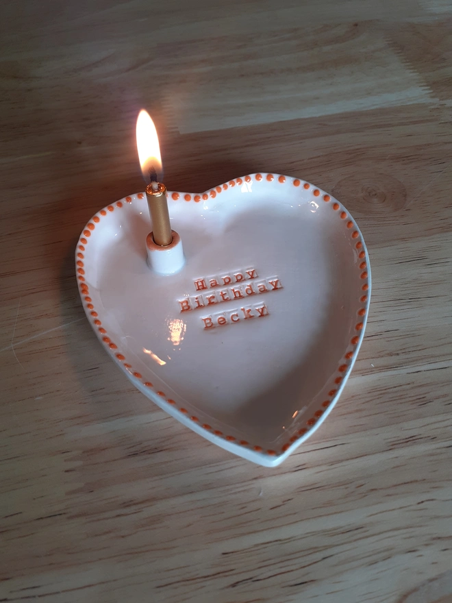Personalised ceramic heart birthday dish shown with the gold candle lit in the integrated holder, highlighting the stamped name “Becky” and the warm glow of the flame.
