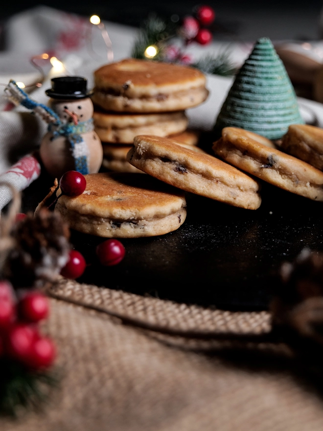 Mince Pie Welsh Cakes displayed on a black bakestone on a busy christmas themed kitchen work top, surrounded by christmas decorations