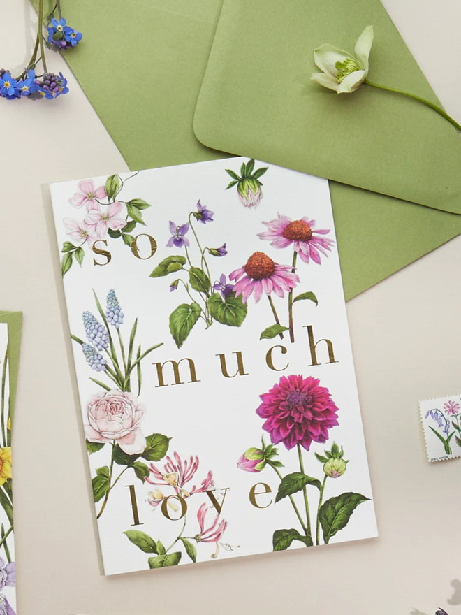'so much love' illustrated floral card. a white floral card and green envelope photographed against a plain background. 
