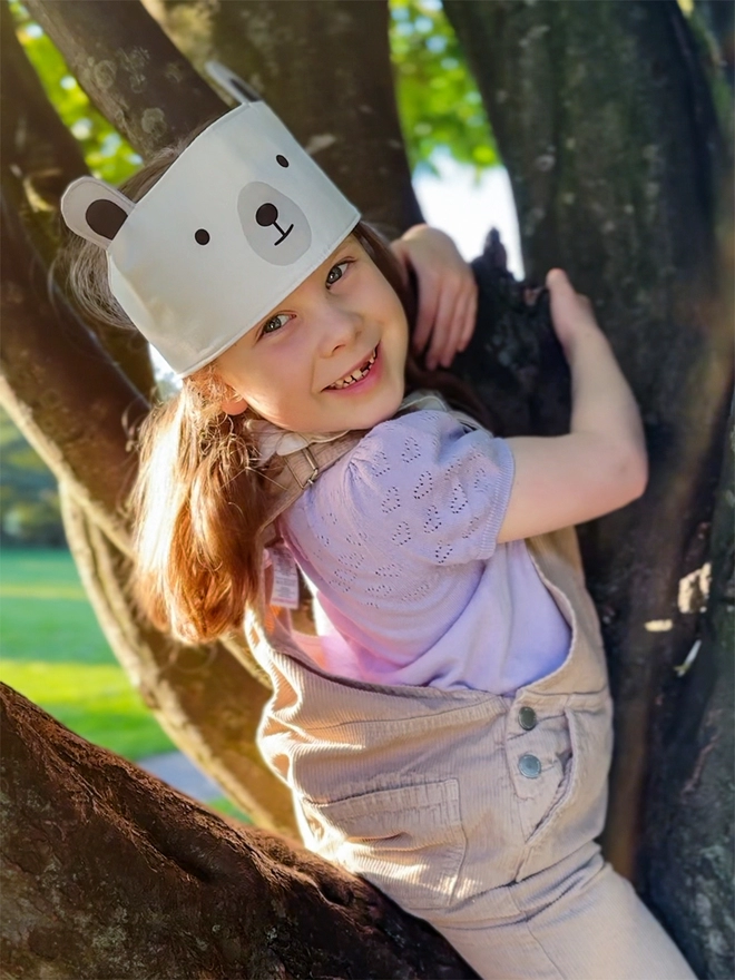 A young girl wearing a dress-up polar bear fabric crown and beige dungarees is climbing a tree.