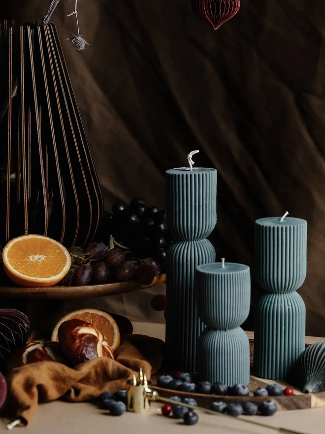 Forest Green Christmas Cracker Candles (Set Of 3). Teal ribbed hourglass candles beside a fruit stand of oranges and grapes, with bread rolls, a linen napkin and a slatted wooden vase. The rich colour and textured design bring warmth and sophistication to winter dining tables.