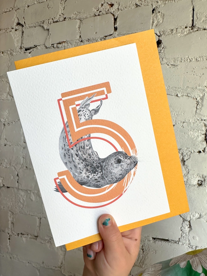 A white card featuring a big orange number 5 and a harbour seal illustration with a bright yellow envelope