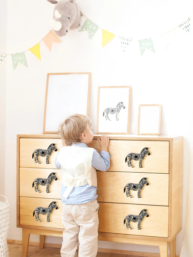 A 4 year old boy leans against a wooden dresser looking at a picture of a zebra, there are six zebra drawer knobs on the dresser, three on each side of him, there is a stuffed to rhinocerous on the wall and bunting.