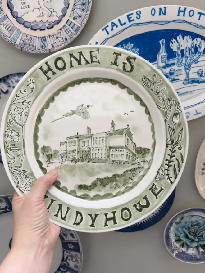 Green plate held aloft by a hand. A pheasant flies over a large house on a hill, with the words "Home is Windyhowe" around the edge, with a botanical border