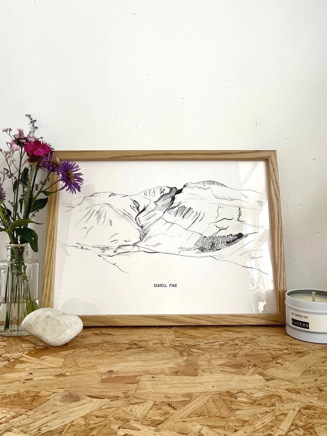 A print of a line drawing of Scafell Pike Mountain in the Lake District, in a frame next to some flowers, a rock and a candle