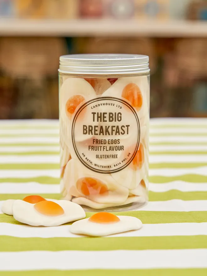 Clear jar of giant fried egg sweets with a silver lid on a green and white striped tablecloth in a garden setting - there are a few fried eggs scattered on the table