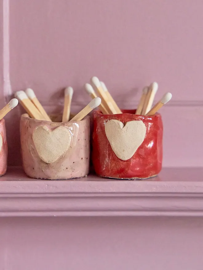 Mini ceramic match holder in red and pink on a pink shelf with white matches inside.