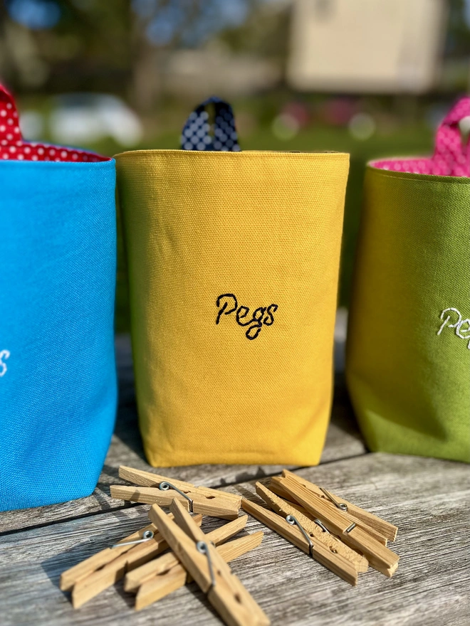 Yellow cotton peg bag with navy polka dot lining and hanging loop, embroidered with 'Pegs', standing on a garden bench with clothes pegs.