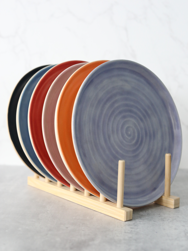 Six handmade ceramic lunch plates displayed upright in a natural wood plate rack, fanned out to reveal a colour palette of pacific blue, mandarin orange, blush pink, cherry red, amethyst purple, and black, against a white marble background