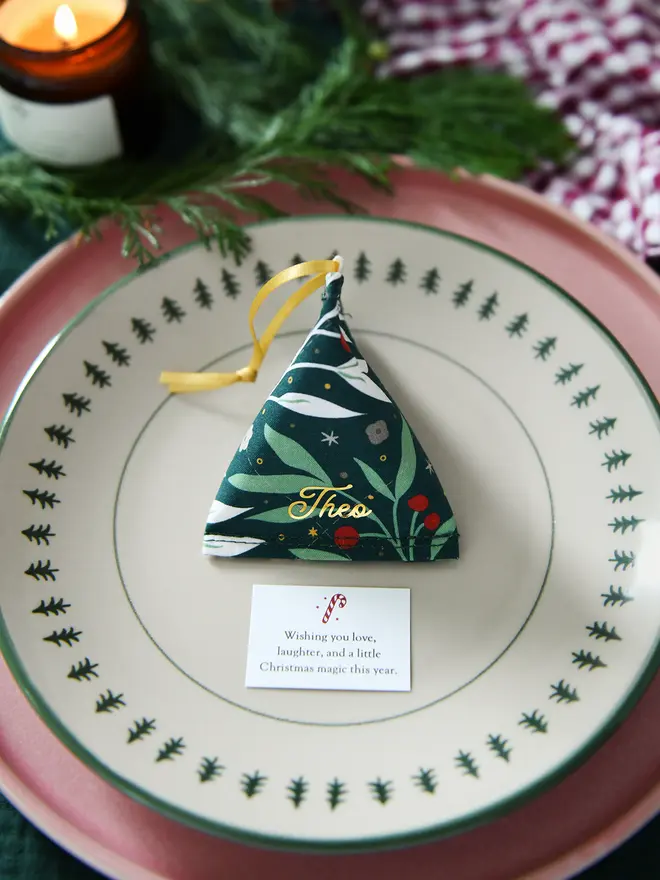 A green festive foliage fabric fortune cookie with gold personalised name “Theo” sits on a Christmas dinner plate, ready as a handmade festive place setting with a hidden Christmas wish card beneath.