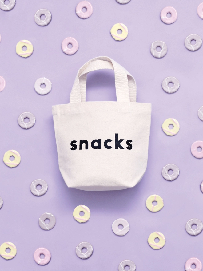 a mini kid's size tote bag with the word snacks on it laying on a purple backdrop with biscuits surrounding it