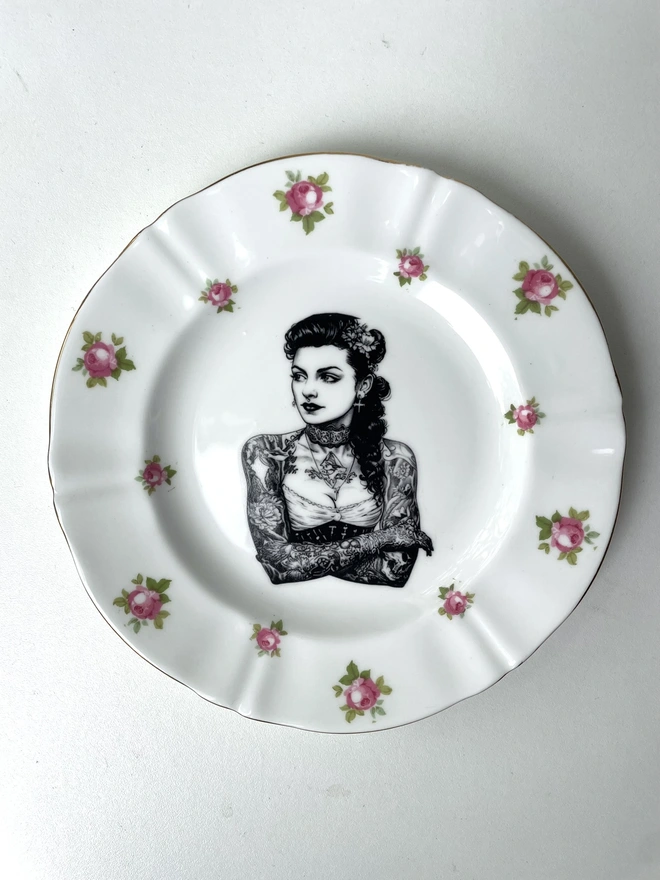a beautifully ornate plate and in the centre is a vintage image of a beautiful tattooed london lady, she has ringlets and a choker and a tattooed ship on her chest
