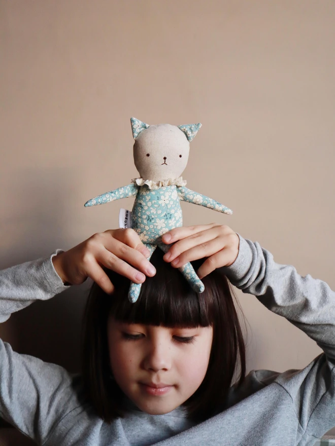 A soft fabric dolls, shaped like a cat with pointy ears and a floral outfit in blue and white lace ruffled collar with simple stitched facial features and a small “BOKUNO” tag on the side.