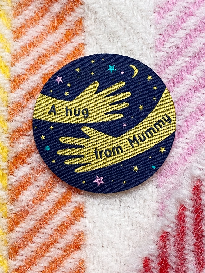 A navy blue and yellow woven patch is laying on a rainbow blanket. The design is two arms in a hug shape with the words "A hug from Mummy" along them. 