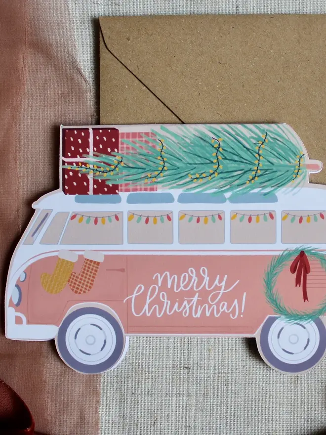 merry christmas illustrated campervan card. campervan shaped card emblazoned with merry christmas and carrying a christmas tree on its roof.