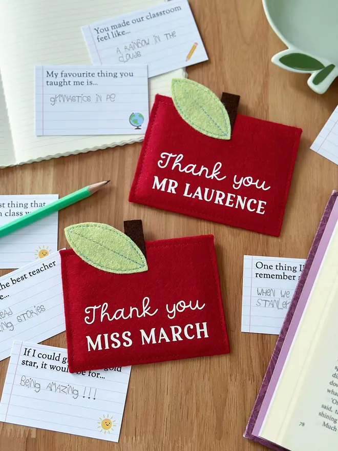 Two handmade red felt apple-shaped envelopes with green felt leaves and brown stems, each reading "Thank you MR LAURENCE" and "Thank you MISS MARCH", surrounded by illustrated prompt cards with child-friendly questions, displayed on a wooden desk with a pencil, notebook, and book. It's a charming personalised, handmade teacher thank you gift.