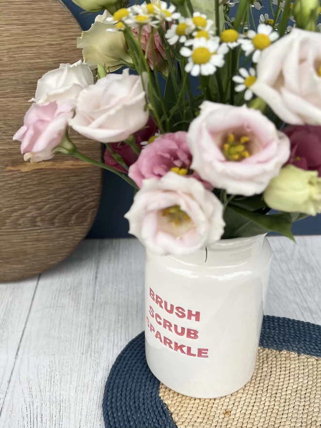 A handmade ceramic ‘Brush Scrub Sparkle’ Pot (lettering in Red) is being use to display flowers.
