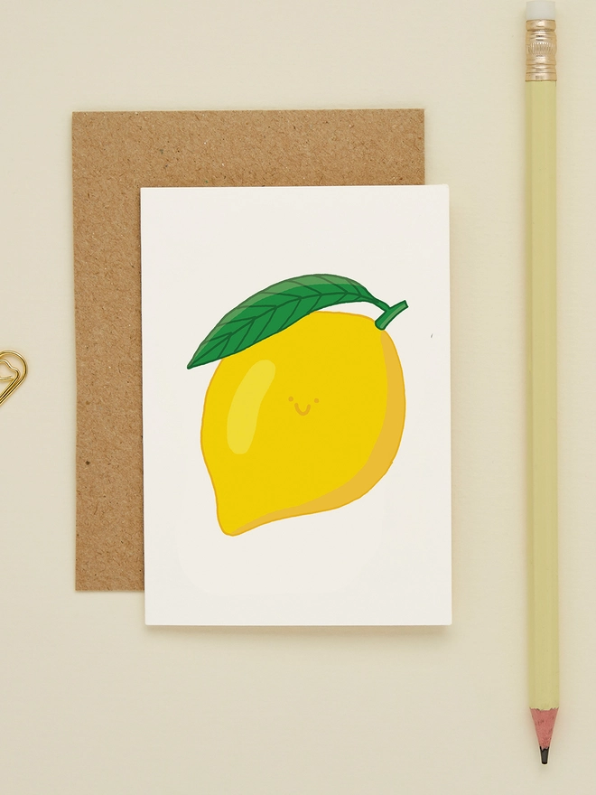 Close-up of miniature lemon card on cream background, paired with kraft envelope and yellow pencil, showing cheerful fruit design
