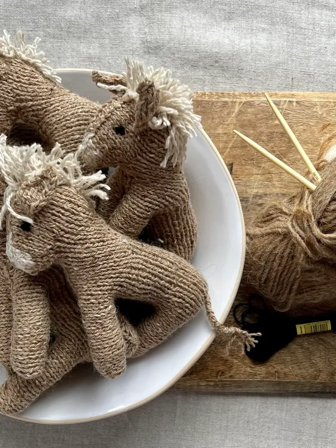 Hand Knitted Natural Dyed Wool And Alpaca Yarn Donkey Gift, a group of hand knitted donkeys sitting in a white bowl on a wooden chopping board on a table cloth. They are accompanied by some hey and some knitting accessories. 