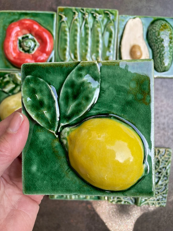 Lemon tile - square, 3D, realistic and glossy. The bright zesty lemon is set off by the lush green glaze of the background. Other fruit and vegetable tiles in the series are on display in the background: fig, savoy cabbage, red capsicum pepper, pear, mange tout, avocado, garlic, asparagus.