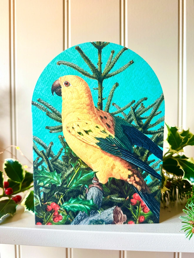 A photo showing an arched shaped Christmas card with a yellow parrot on a Christmas tree with holly. The card sits on a shelf, displayed with Christmas foliage.