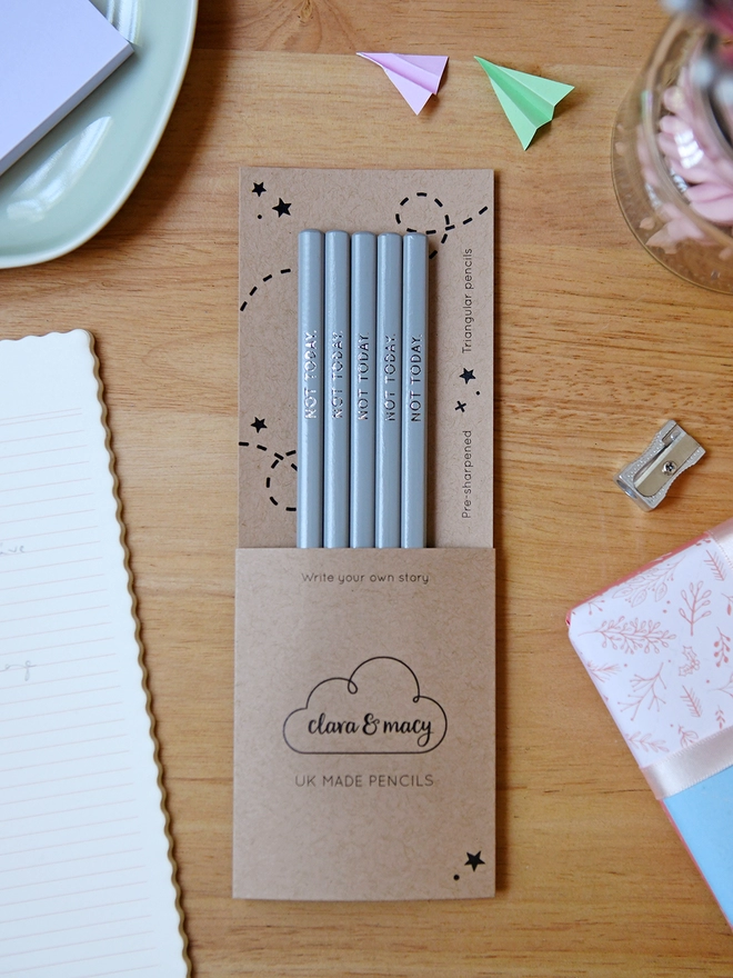 Five grey pencils with the words Not Today written along the side, sit within cardboard packaging on a wooden desk with stationery items.