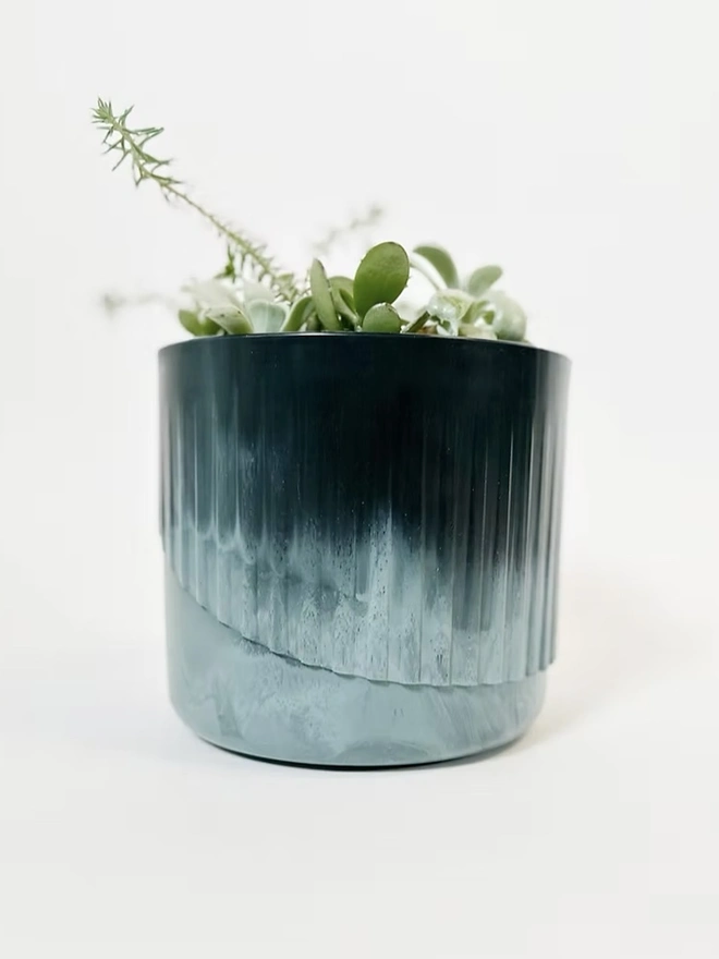 Ocean Plastic Plant Pot Medium, a recycled blue plant pot sitting on a plain surface against a white backdrop. It is accompanied by some plants. 