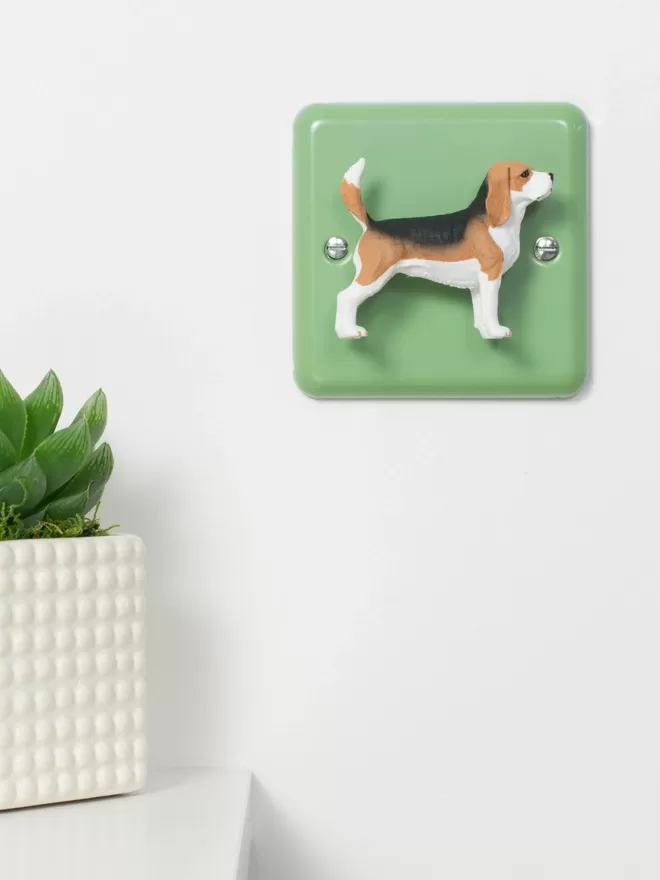  A beagle dog dimmer light switch. The light switch has a pastel green steel metal epoxy coated light switch plate, the beagle is very lifelike and made of plastic. The light switch is brand is Candy Queen Designs and Varilight. The beagle light switch is on a white wall, to the left there is a small succulent plant in a square white vase.
