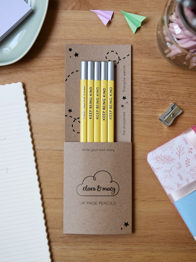 Five yellow pencils with the words Keep Being Kind along the side of each one, are tucked into cardboard packaging on a wooden desk.