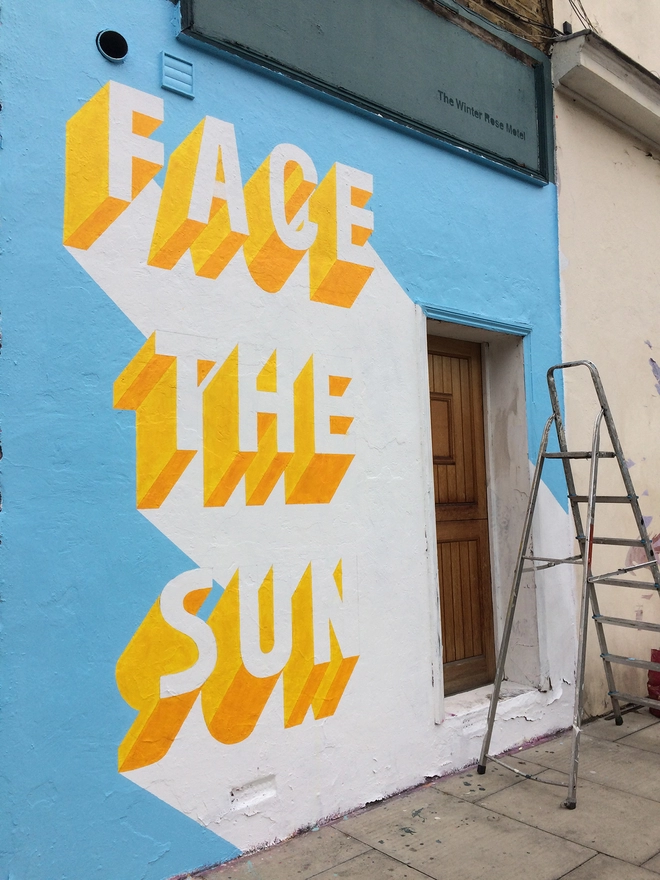 A Survival Techniques street art painting in progress with 3d typography of the words Face The Sun and a ladder