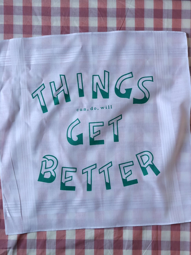 A Mr.PS Things Get Better Handkerchief printed in green, laid on a pink gingham tablecloth