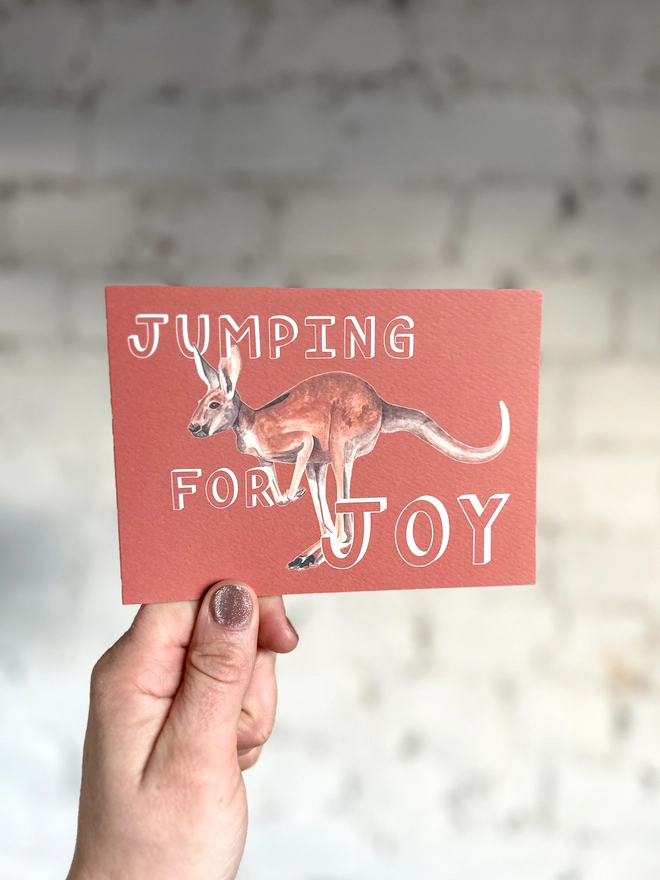 a rusty red greetings card featuring an illustration of a kangaroo with the text “jumping for joy”