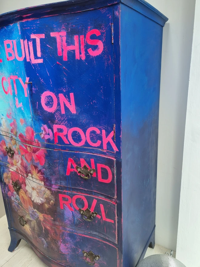 A vintage tallboy cabinet painted in vibrant blues, neons and flowers with the text that reads 'we built this city on rock and roll' in neon pink.