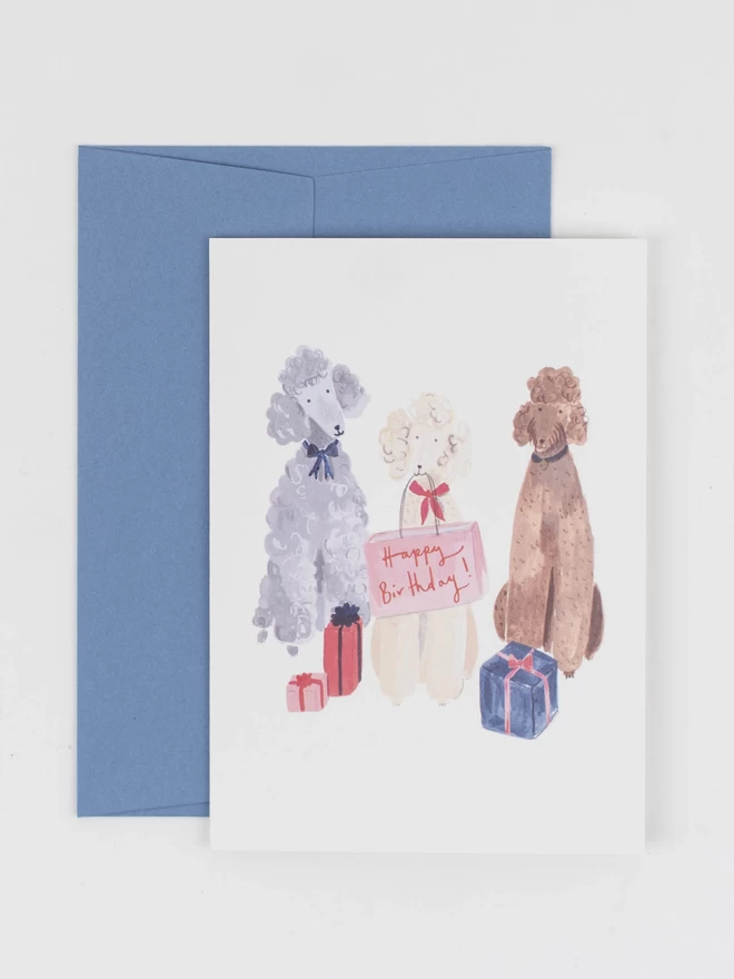 An illustrated greetings card, with corresponding pale blue envelope. The card features three fluffy poodles surrounded by gifts and holding a shopping bag that reads "happy birthday"