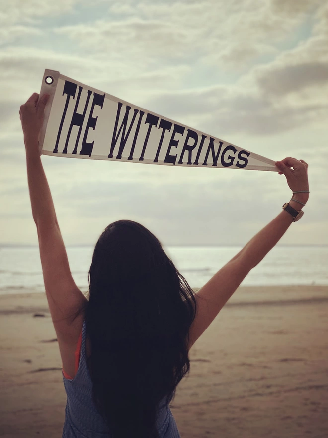 Someone at the beach holding a pennant flag in the air. The pennant is ivory with navy writing saying The Witterings