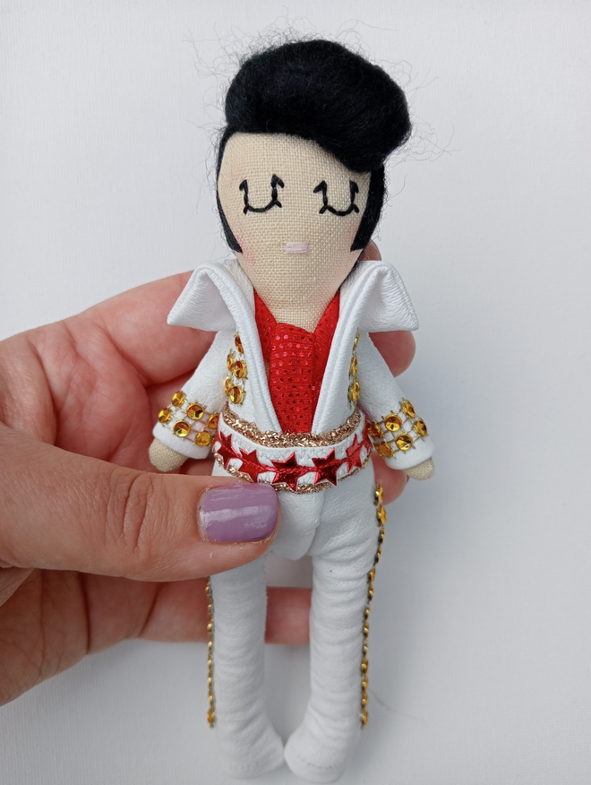 Mini decorative Elvis Presley icon doll held in a left hand for scale Elvis is wearing a white jumpsuit adorned with gold sparkles and a red metallic star belt