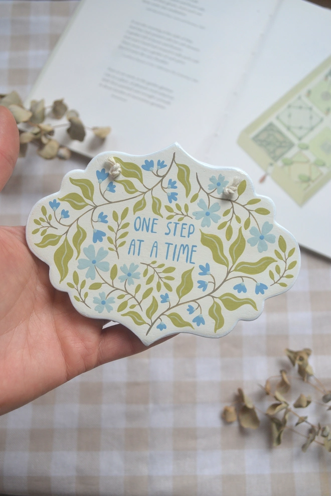 hanging clay sign with blue and green floral design reading 'one step at a time' in front of gingham background