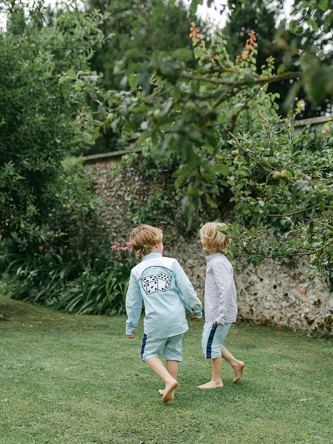 Two little boys walk through a garden. One wears a cowboy shirt with dice appliqued on the back.