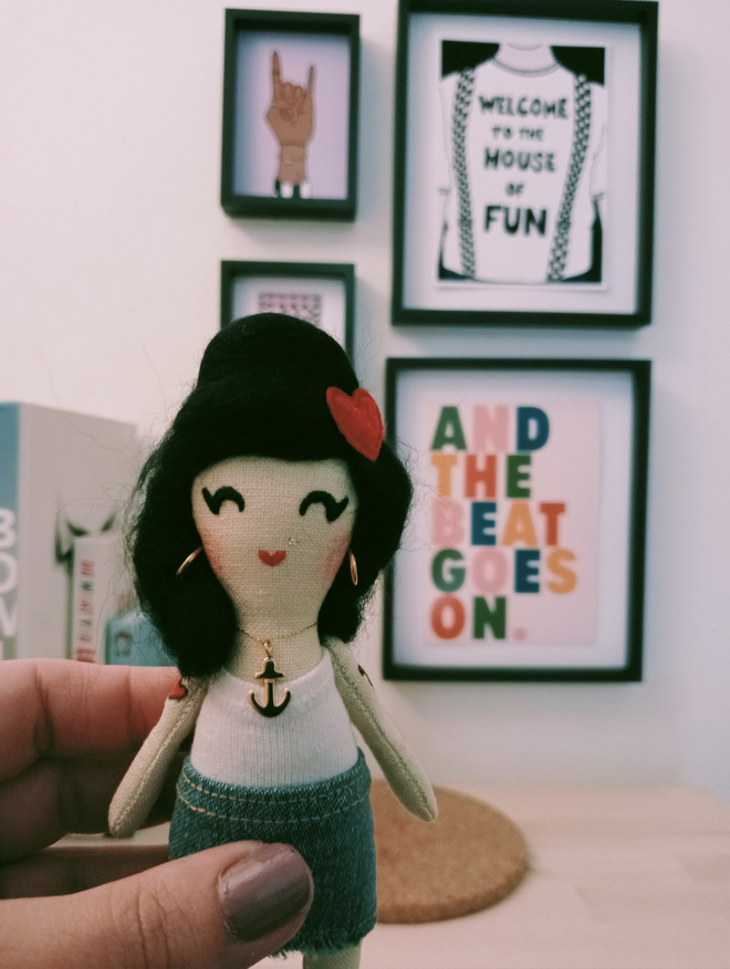 Amy Winehouse mini decorative doll held up to camera in left hand wearing a beehive gold hoop earrings and anchor charm with red love heart hair clip