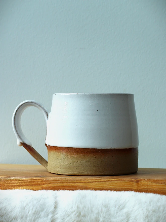 Handmade ceramic mug with a glossy white upper glaze and a natural toasted clay base, against a soft green wall background.