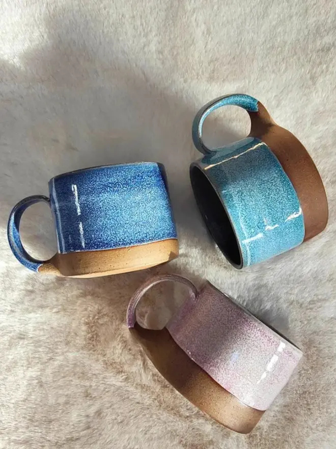 Three handmade ceramic cups with pastel glazes arranged in a circle on a soft cream-colored fur surface.