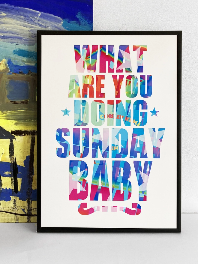 Framed multicoloured typographic print of a Pulp song lyric from Common People - “What are you doing Sunday, baby?”  The print rests against a blue and yellow abstract painting.