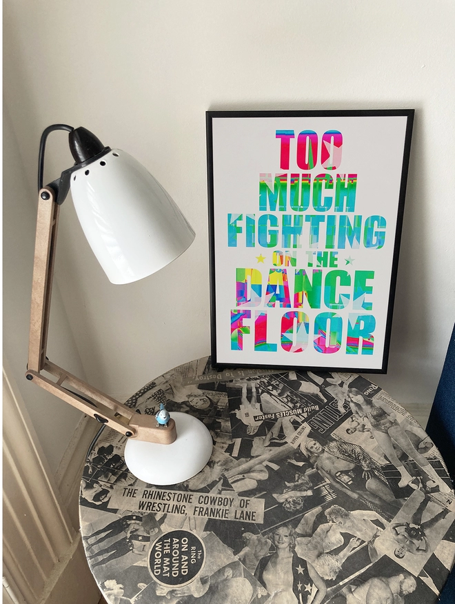 Framed multicoloured typographic print - Lyrics from The Specials song Ghost Town "too much fighting on the dance floor" . The print sits on a round table decoupaged with 1970's wrestlers next to an anglepoise lamp.