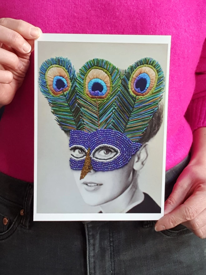 Black and white photograph print, woman wearing embroidered peacock mask held against pink jumper