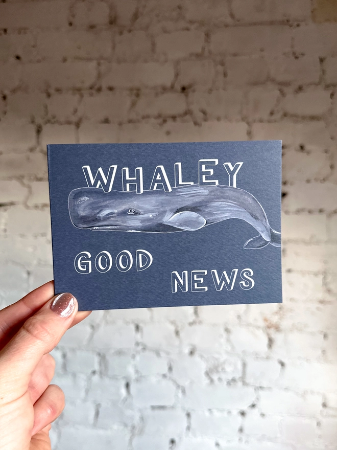 a dark grey blue greetings card featuring an illustration of a sperm whale with the text “whaley good news”