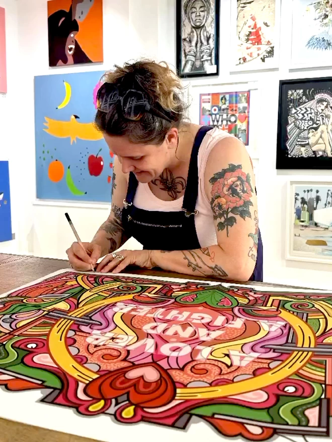 Rebecca Strickson sits signing an “A lover and a fighter” print. She is a white woman wearing a white top and denim dungarees. She has many tattoos, including a rose on her upper arm closest to the camera.