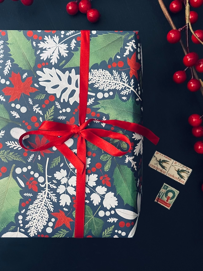 Christmas present wrapped in Winter leaf gift wrap with Holly and Ivy design, red ribbon, red berries, and Xmas postage stamps on dark table