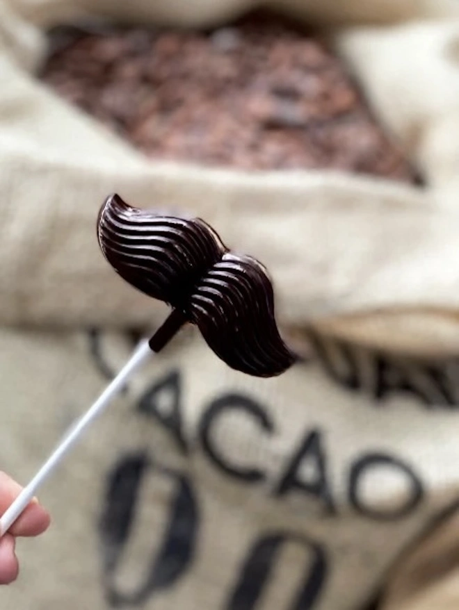 Moustache Chocolate Lolly 58%, a person holding a chocolate lolly in front of a bag of nuts. 